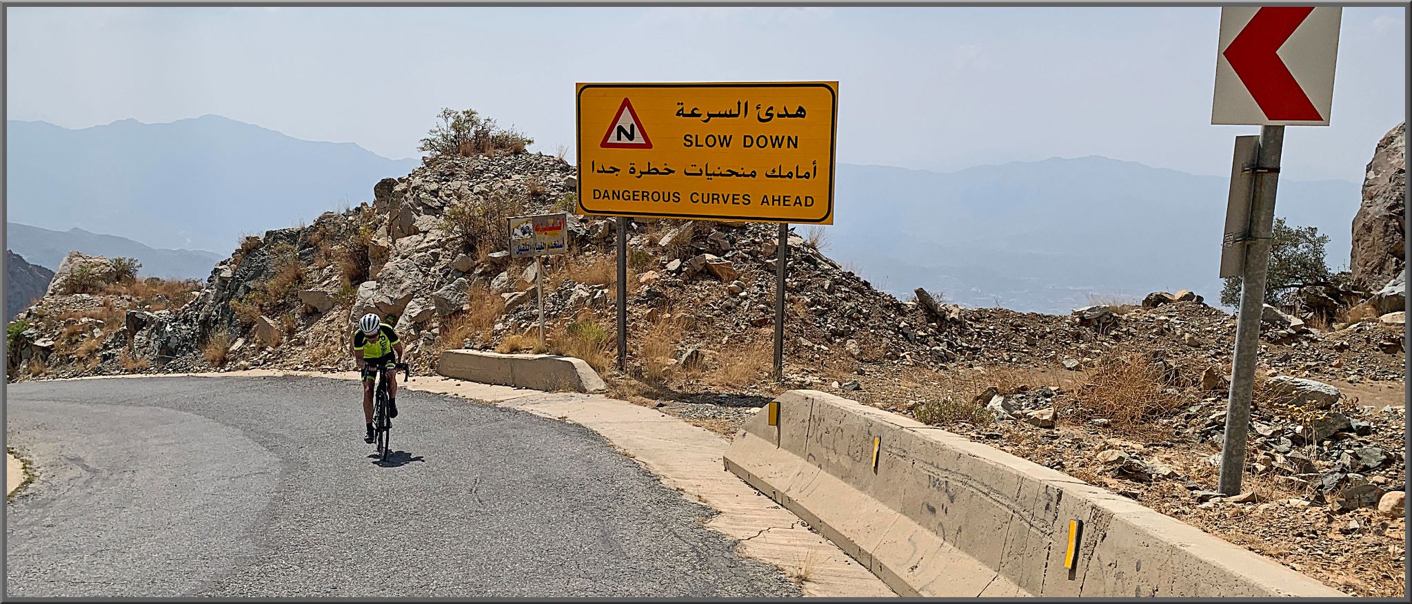 Cycling Sinan Road (Al Meger), Saudi Arabia - PJAMM Cyclist rides up roadway near roadsign that warns, "Slow Down: Dangerous Curves Ahead"