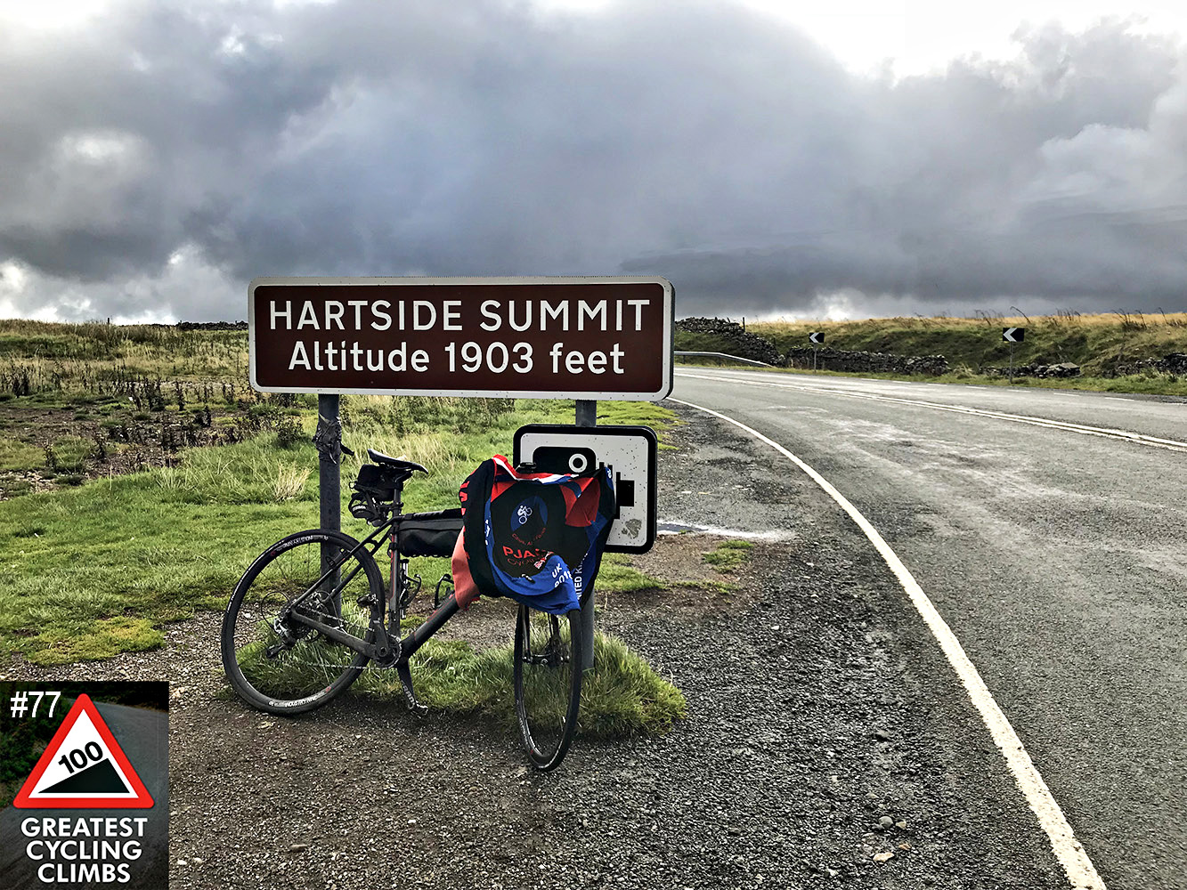 Cycling Hartside, UK - Simon Warren's 100 Greatest Cycling Climbs #77 logo in corner, road bike leaning against road sign for Hartside Summit Altitude 1903 feet, grey skies