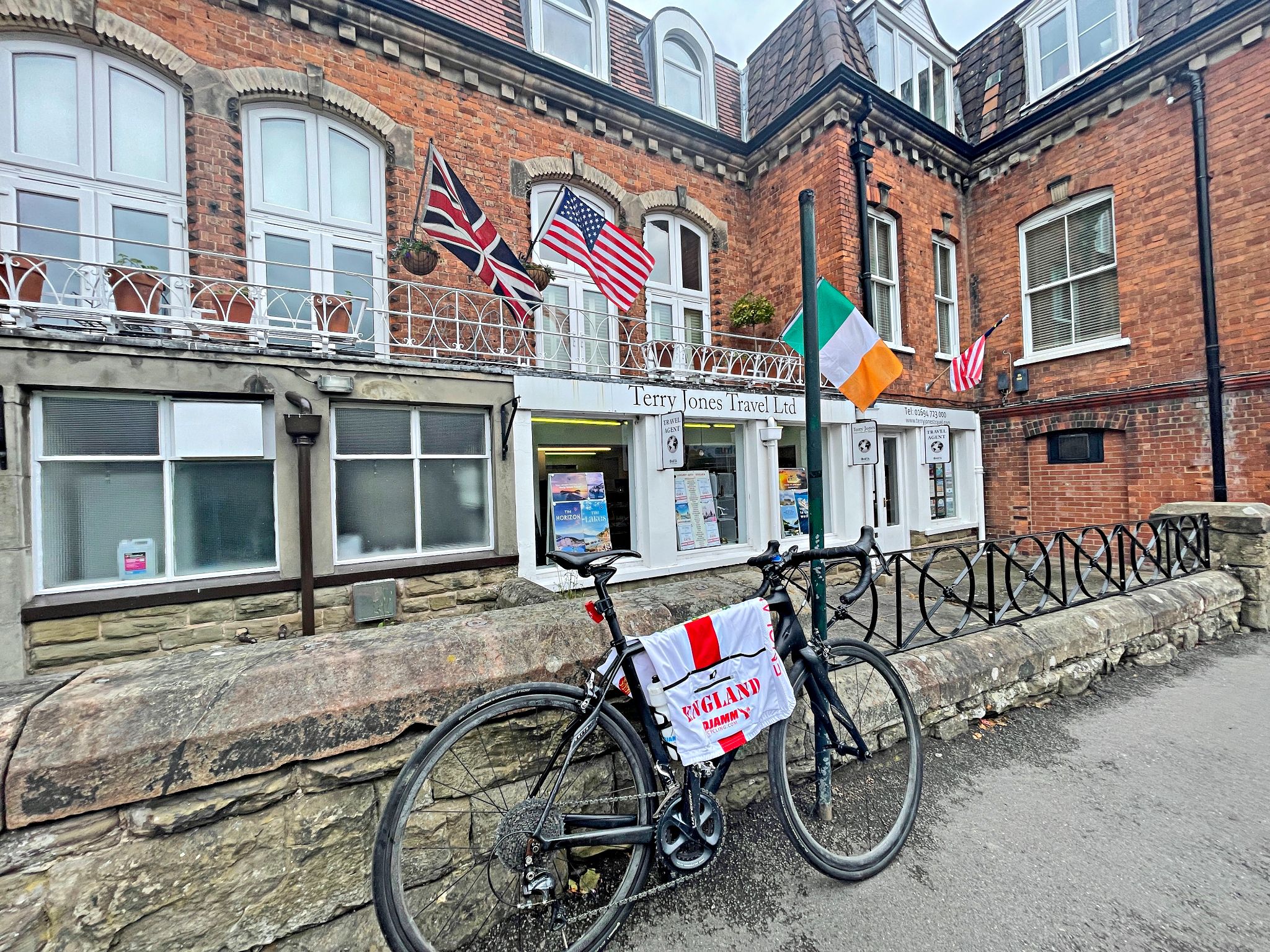 Cycling The Burway, Church Stretton, Wales bike parked in front of red brick building with flags of UK, USA, and Italy flying