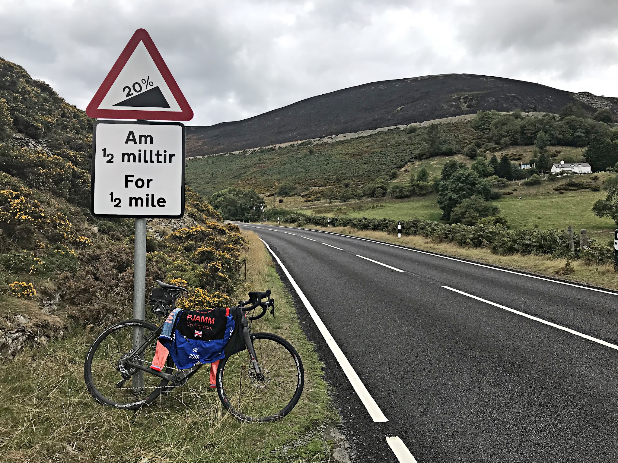 Climbing Horseshoe Pass by bike - bike parked along two-lane roadway leaning against road sign for 20% grade, PJAMM cycling jersey draped across bike, green rolling hillsides and grey cloudy skies