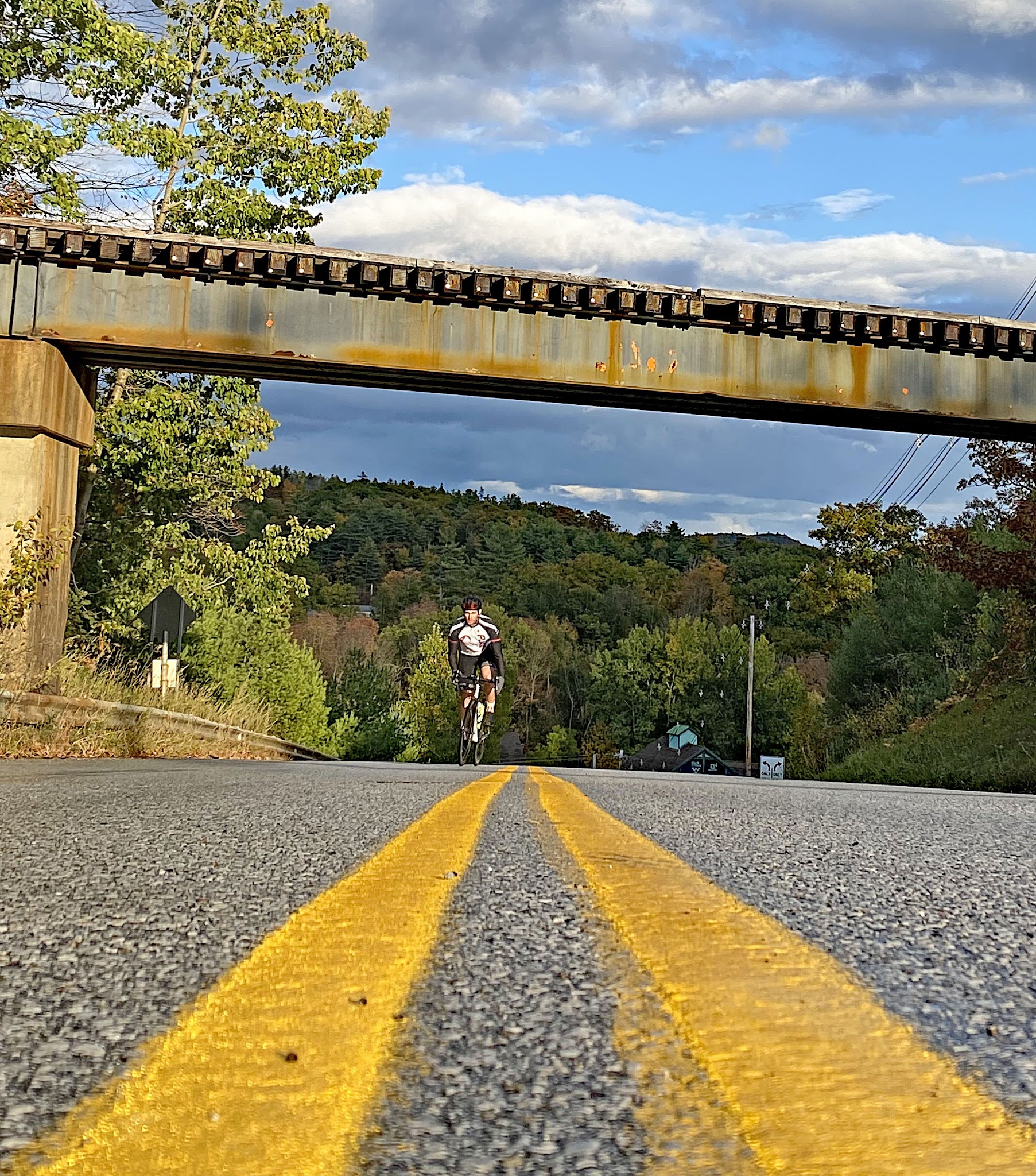 Cycling Okemo, Vermont - cyclist rides on center of two-lane roadway, underneath rustic train bridge crossing roadway