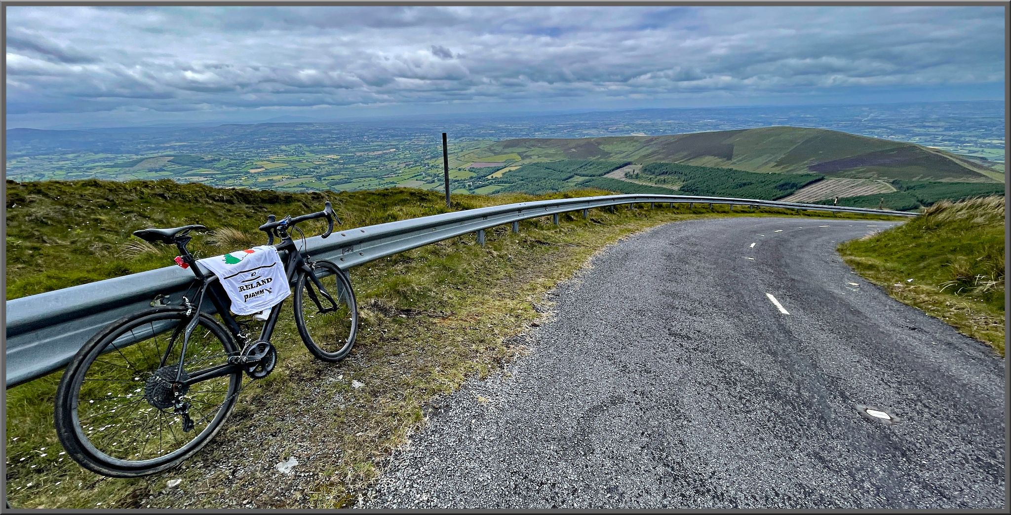 bike parked against guardrail looking down over Irish farmland below; two-lane roadway