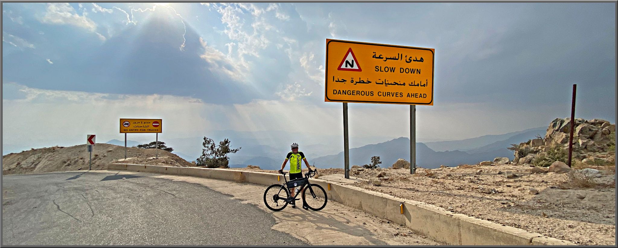 Cycling Almdan Pass, Saudi Arabia - PJAMM Cyclist John Johnson stands with bike on side of road, next to large yellow roadsign reading, "Dangerous Curves Ahead," sun rays beam down through clouds in skyline behind