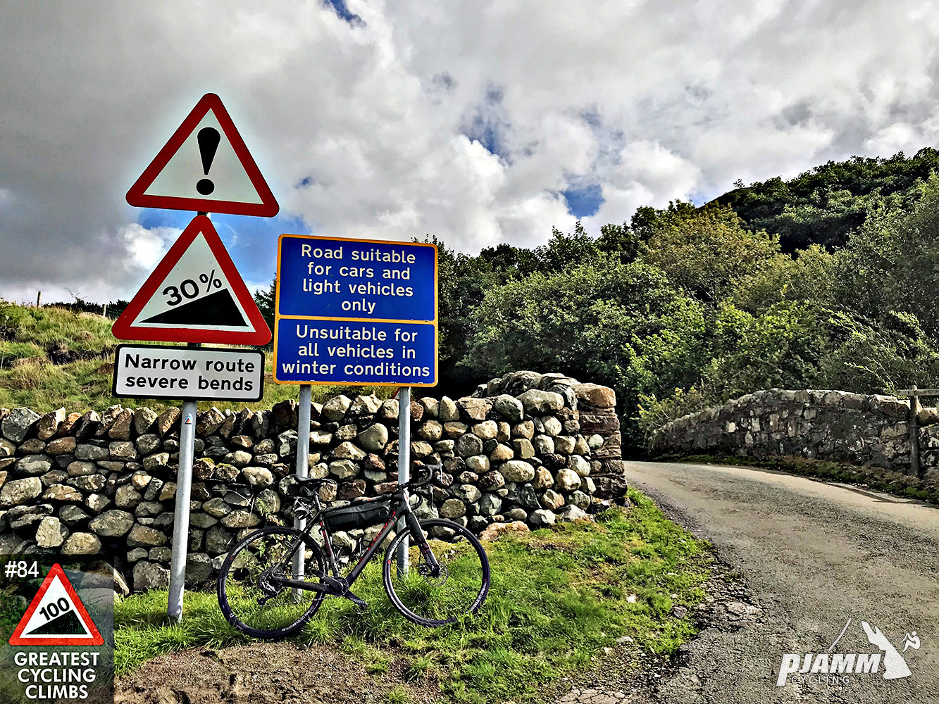 Cycling Hardknott Pass #84 Simon Warren 100 Greatest Cycling Climbs  - bike leaning against 30% steep grade sign, rock wall, roadway, 100 Greatest Cycling Climbs logo