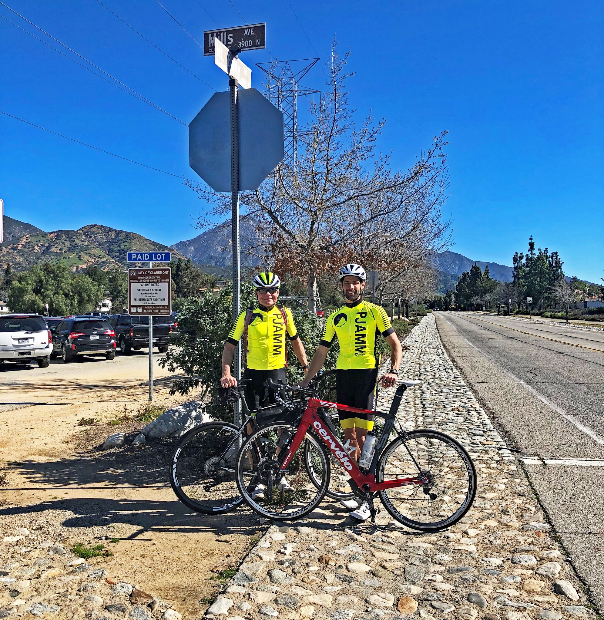 Two PJAMM Cyclists in yellow jerseys stand with their bikes at the intersection of Mt. Baldy Road and N. Mills Avenue