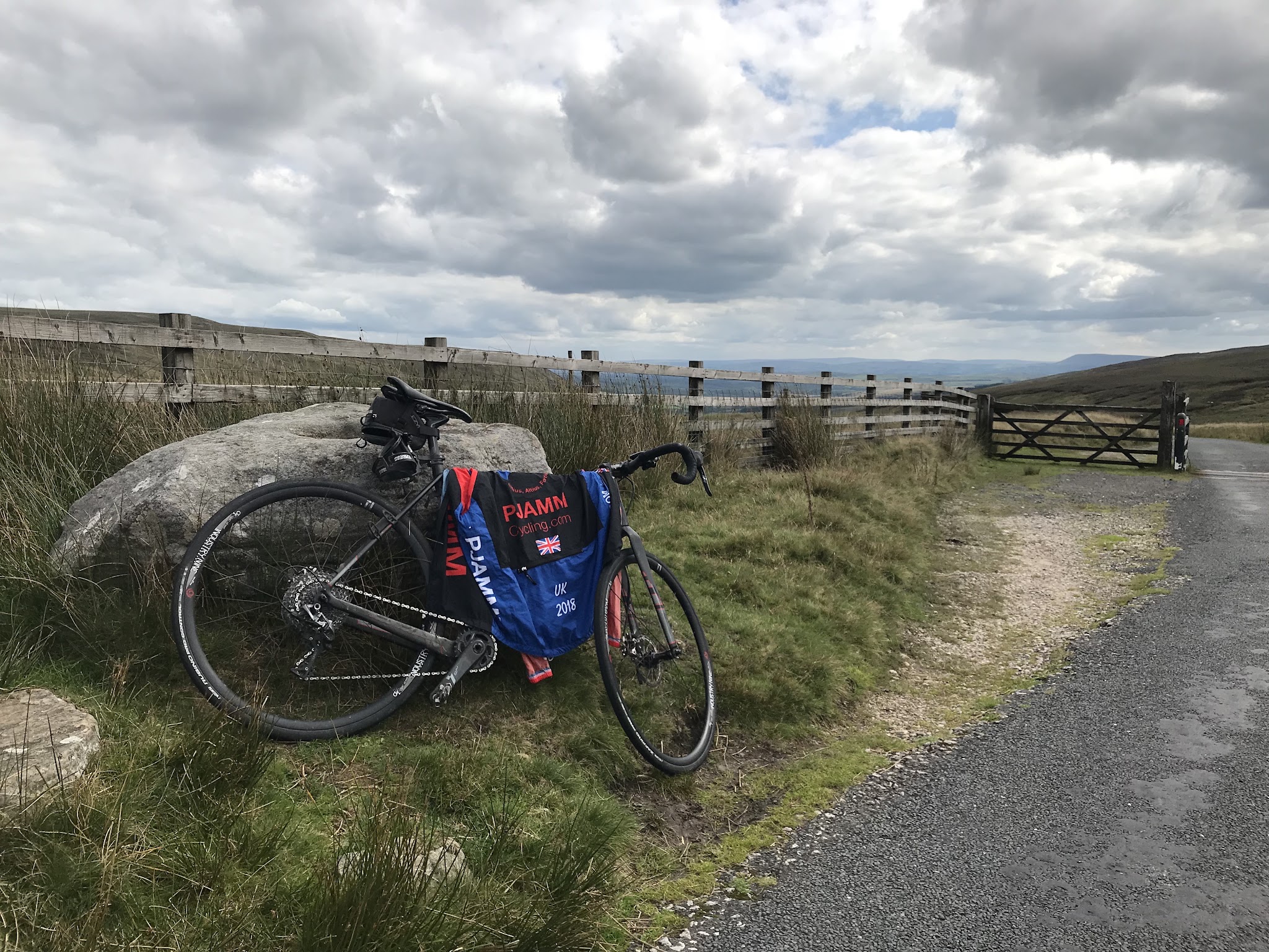 Bicycle climb Cross of Greet #79 Simon Warren 100 Greatest Cycling Climbs  - bike leaning against rock, fence, road, clouds and fields