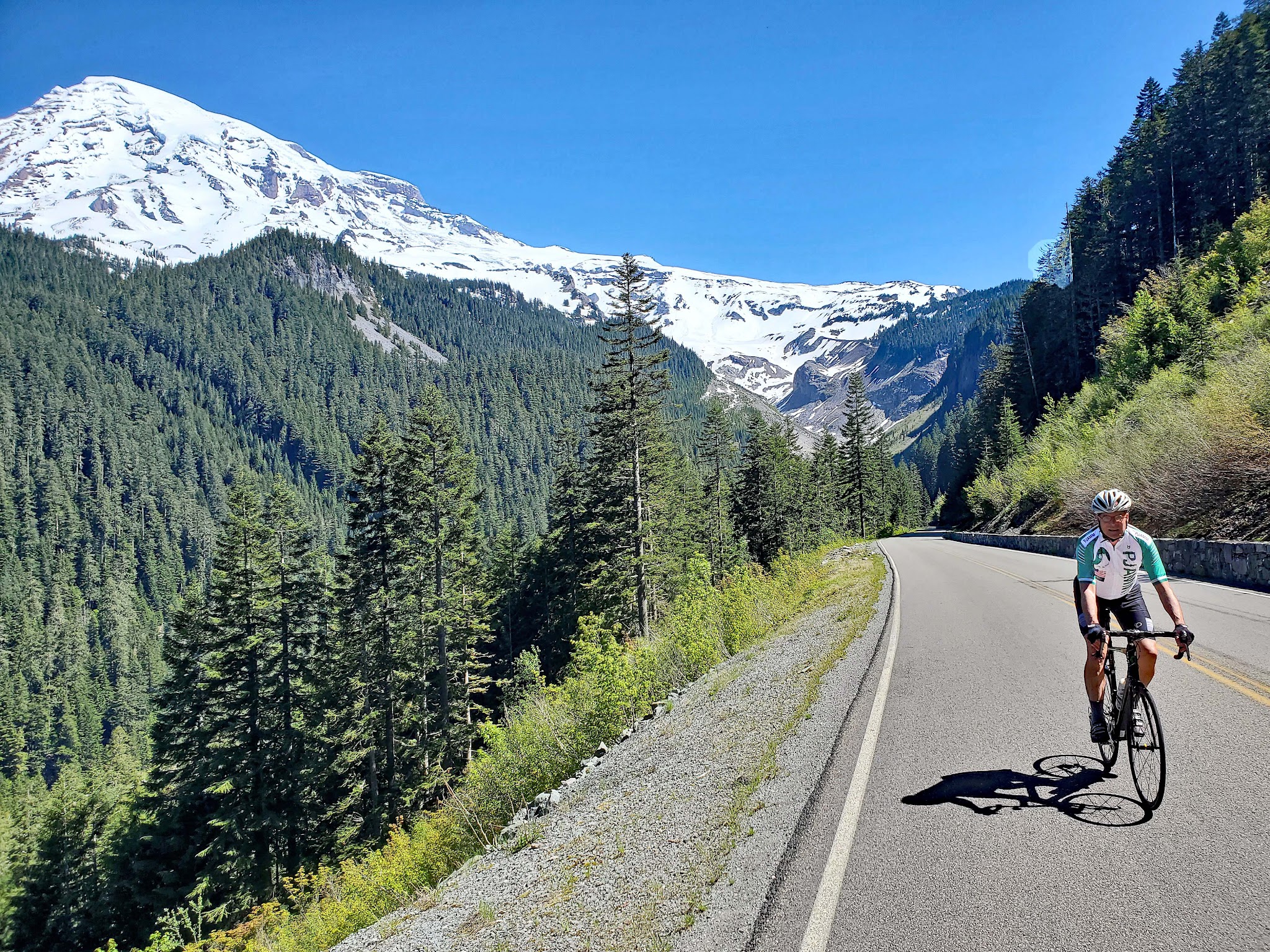 Cycling Mt. Rainier from Nisqually River Entrance - cyclist riding on Paradise Road, evergreen trees grow along mountainside, snow capped mountains in background
