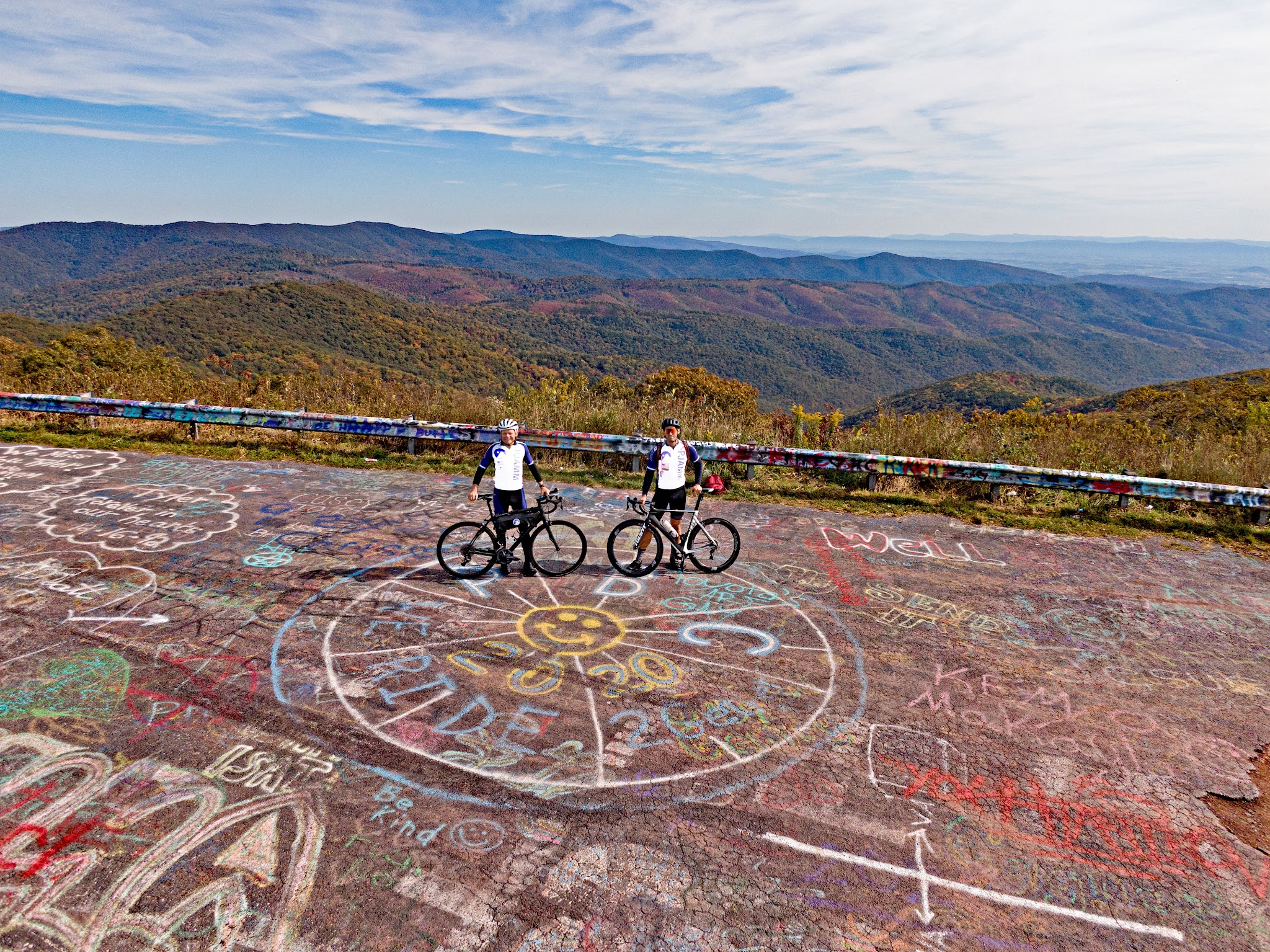 Cycling Reddish Knob, Virginia - aerial view of two PJAMM Cyclists standing at summit, on the border of Virginia and West Virginia, bright graffiti painted all over road, bright fall foliage on hillsides in distance