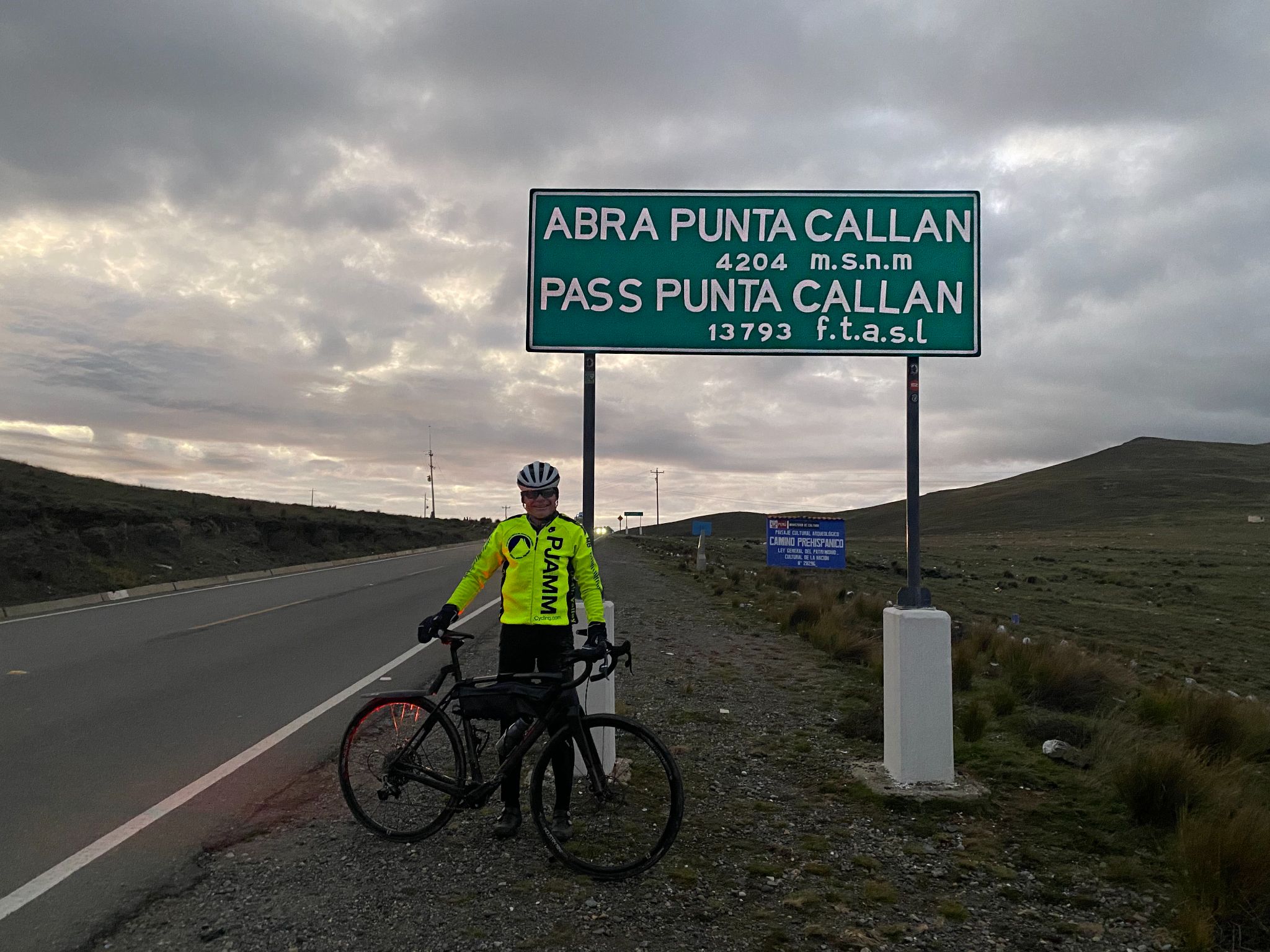 Cycling Punta Callan East, Peru - PJAMM Cyclist John Johnson stands with his bike in front of the green road sign for Punta Callan