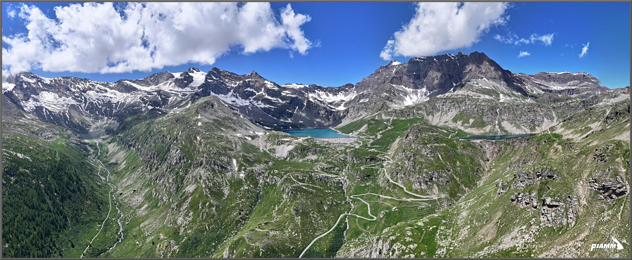 Cycling Colle de Nivolet, Italy - panoramic view shows road snaking up green mountain pass, blue alpine lake at top of mountain