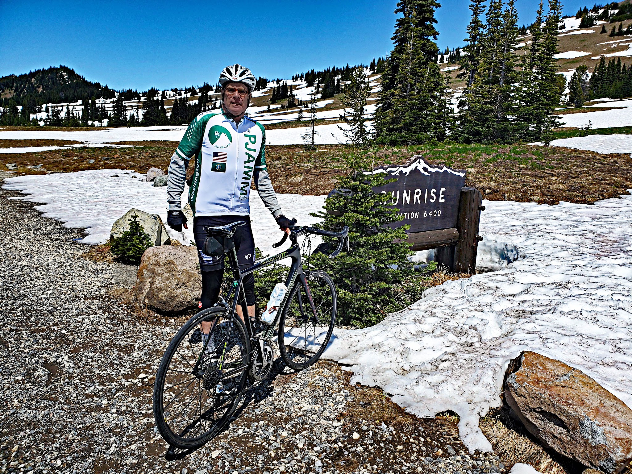 Cycling Mt. Rainier, Sunrise VC, Washington - cyclist wearing PJAMM Cycling jersey standing with bike next to sign for Sunrise, Elevation 6400, snow patches on ground, evergreens in background