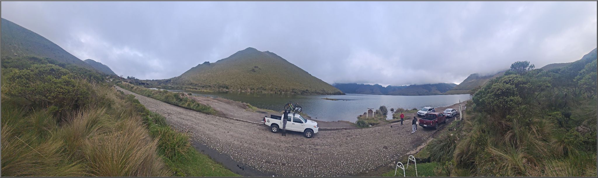 panoramic view of gravel road with white truck being unloaded of bikes, next to large body of water and tall mountain in background