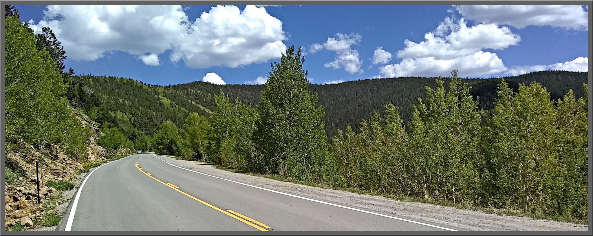 Cycling Juniper Pass East, Colorado - view looking town a straightaway on a two-lane paved road along mountainside, dense forestation and blue sky