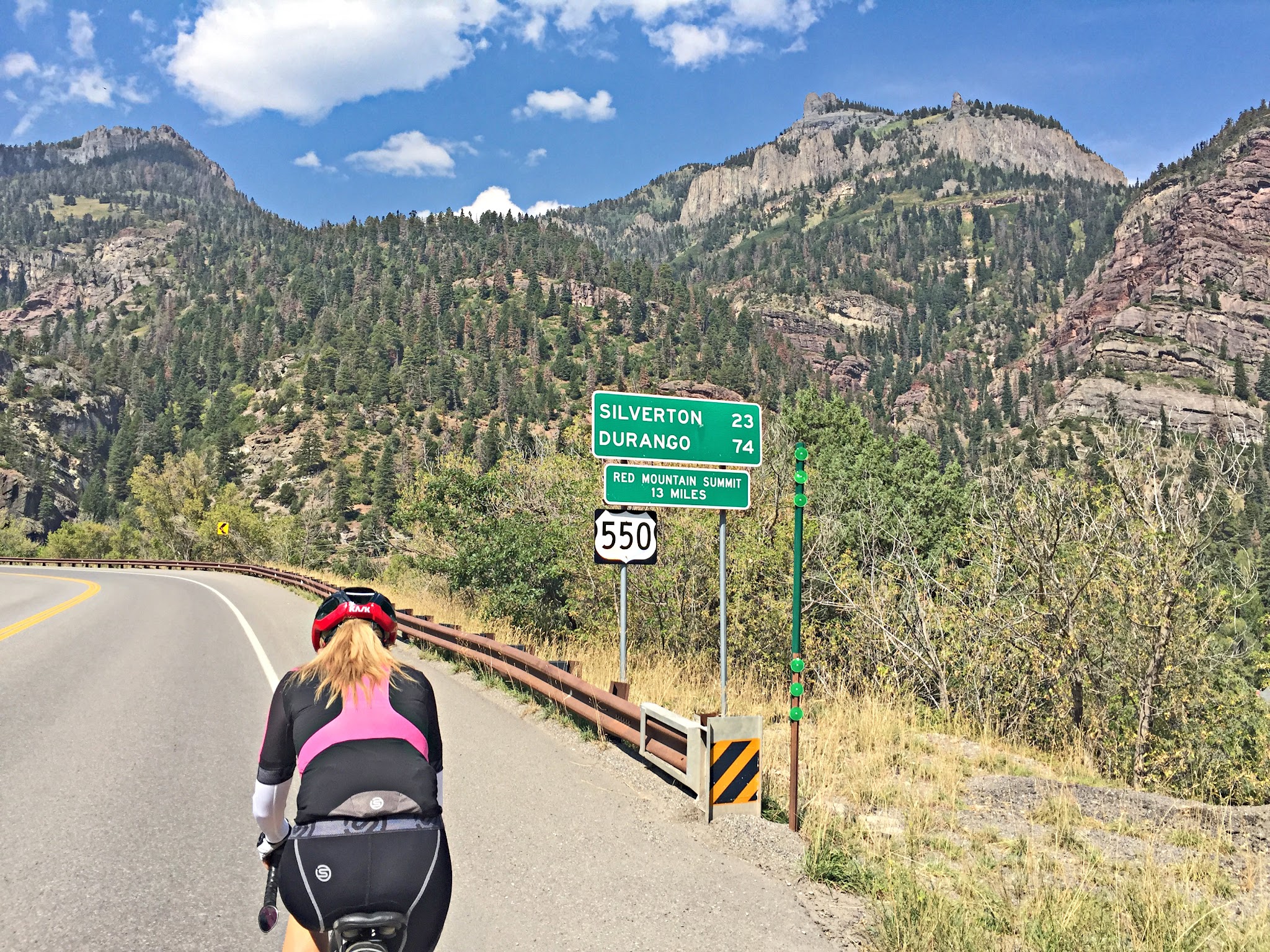 Cycling Red Mountain,  Ouray, Colorado - cyclist riding on highway 550 surrounded by mountains near road sign for Silverton (23 miles away) and Durango (74 miles away)