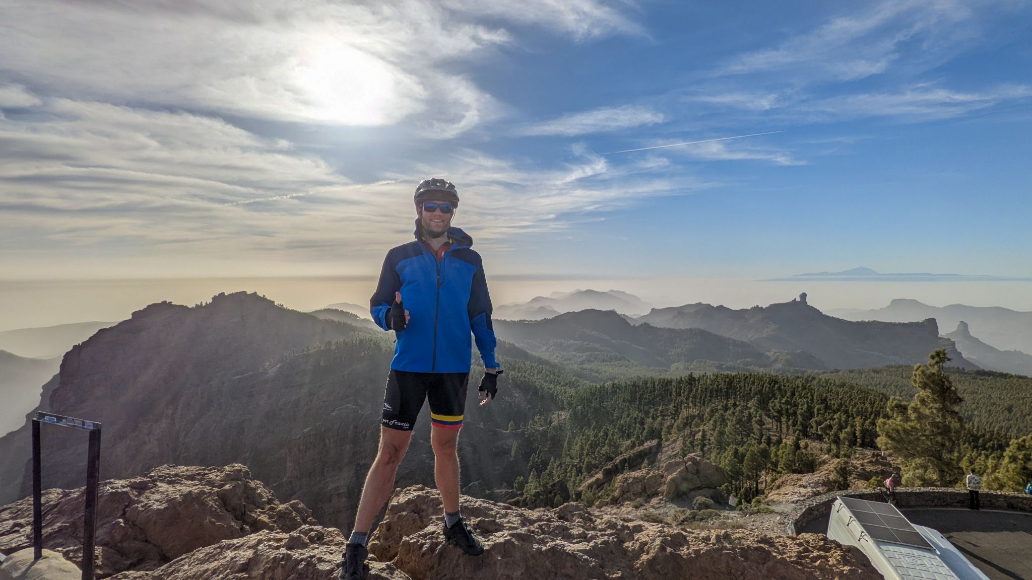 PJAMM cyclist stands on craggy rocks on roadside at climb's finish, beautiful mountain setting with rock formations silhouetted in background