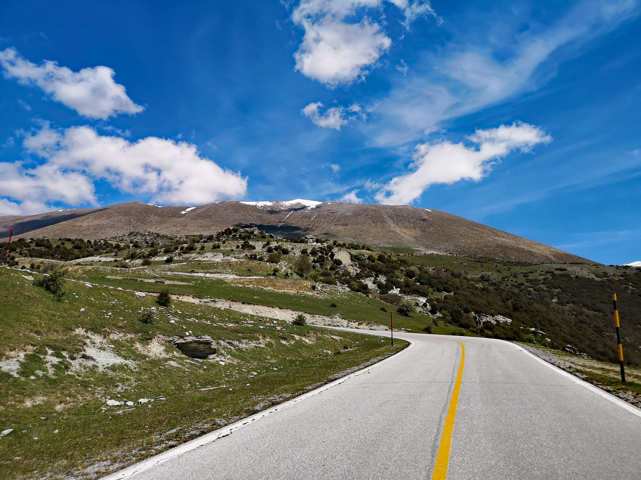 Cycling Mount Olympus, Greece - two-lane roadway headed up mountainside covered in green grass and some shrubbery, blue sky and white clouds beyond