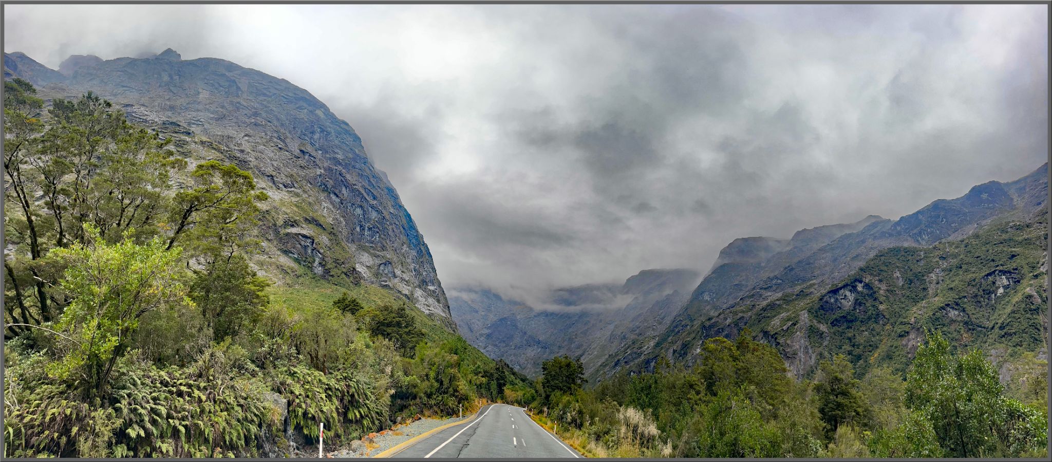 panoramic photo shows straight roadway surrounded by rock mountainsides and thick greenery
