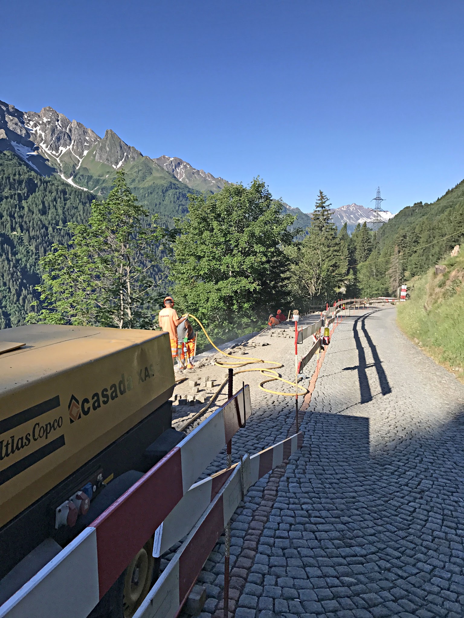 Cycling Gotthard Pass (St. Gotthard Pass), Switzerland - road workers with equipment maintaining cobblestones on historic roadway, Swiss alps in background