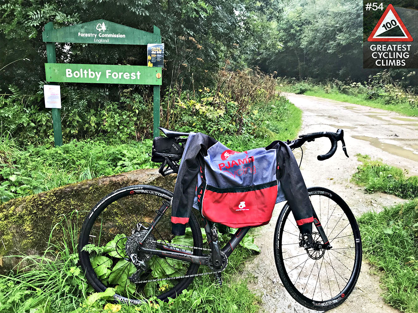 Cycling Boltby Bank, North Yorkshire, England - Simon Warren's Greatest Cycling Climbs #54 logo in corner, bike parked in front of Boltby Forest sign, rainy day, lots of greenery 