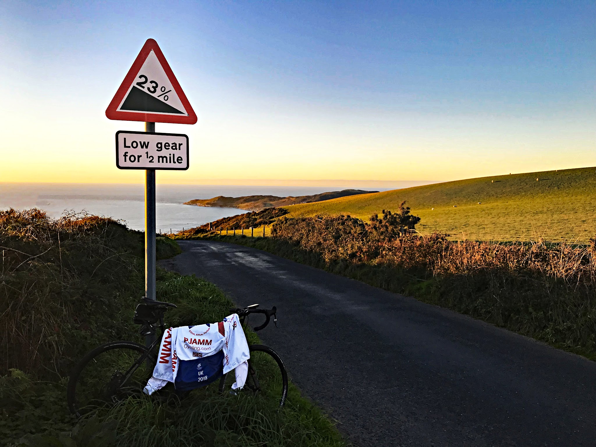 Cycling Challacombe Hill - bike parked against road sign for 23% grade noting "Low gear for 1/2 mile", PJAMM Cycling jersey draped over bike, hillside at golden hour with sun setting over water in background