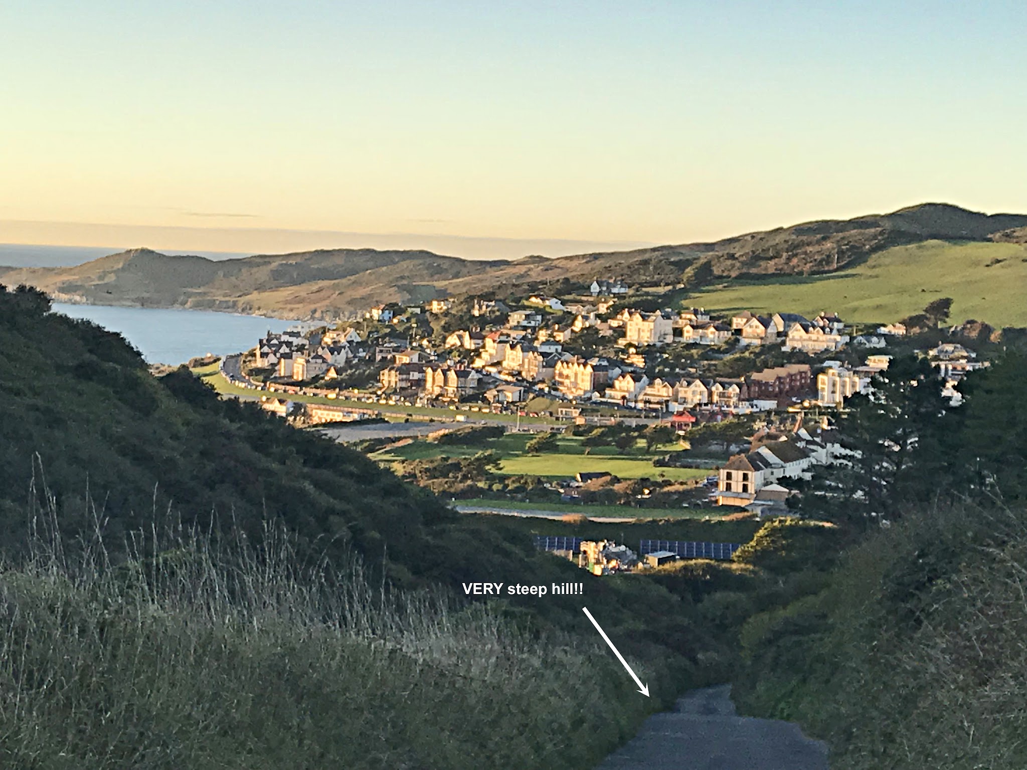 Cycling Challacombe Hill - very steep, very narrow roadway lined with tall grasses, roadway is leading up from a costal village dotted with white buildings and homes, sunset over water in background