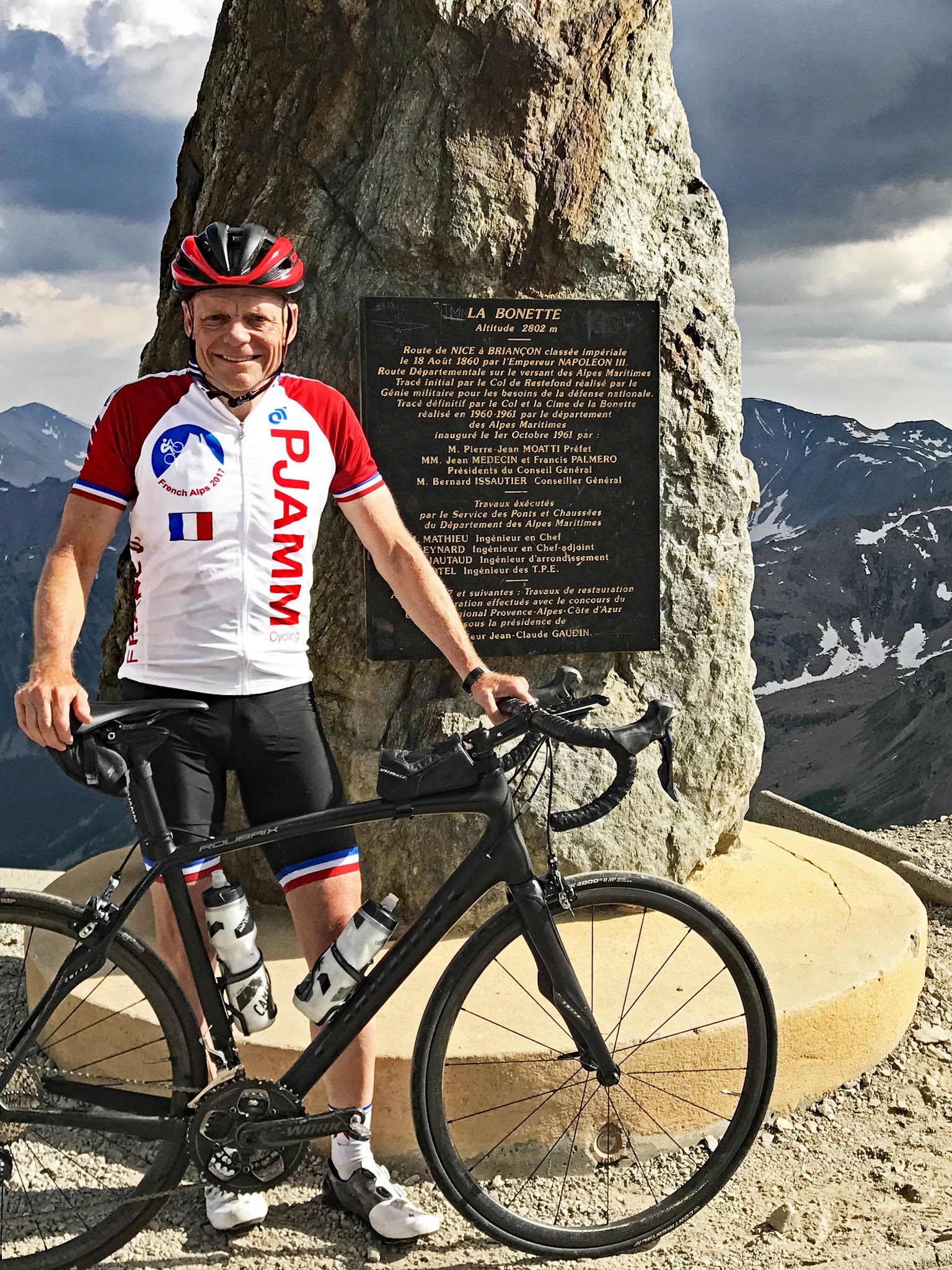 PJAMM Cyclist John Johnson stands with bike at climb's summit in front of stone marker and plaque 