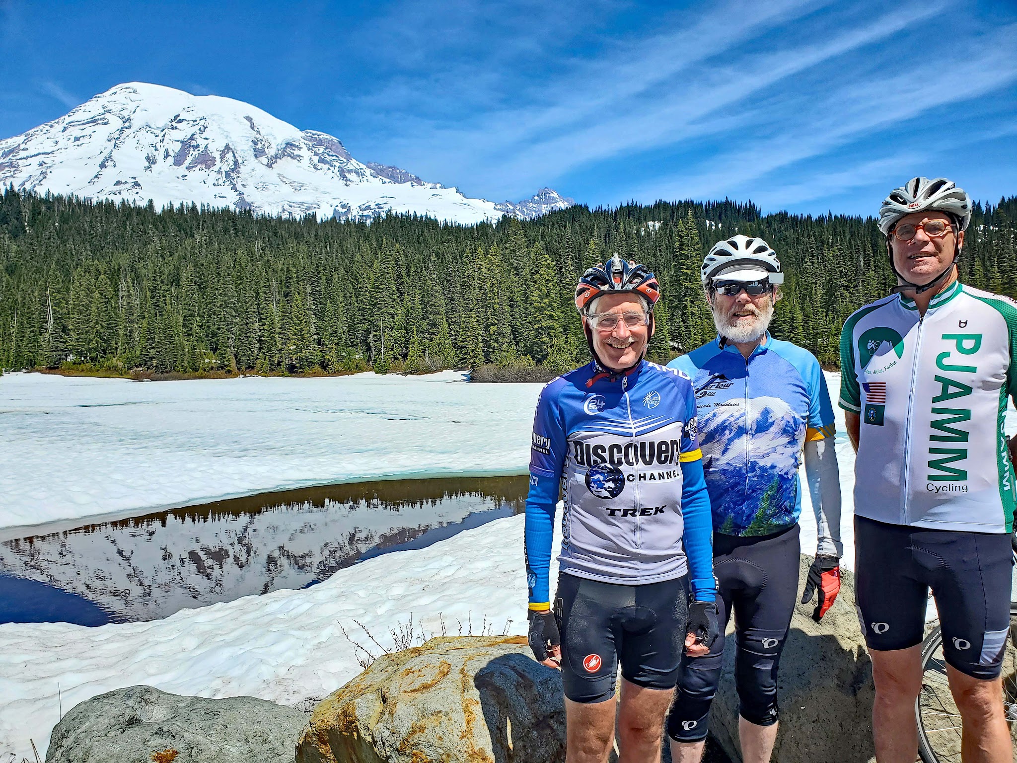 Cycling Mt. Rainier from Nisqually River Entrance - three cyclists standing in front of Reflection Lakes, snow, snow capped mountains, and dense evergreen forestation behind them