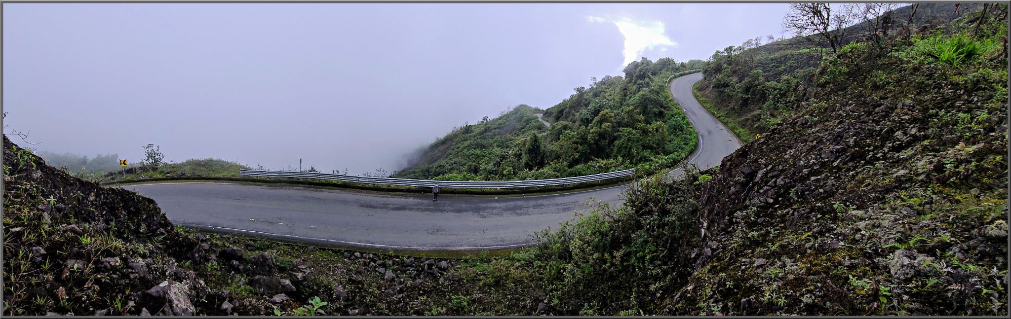 panoramic view of slick, wet, one-lane mountain roadway curving along hillside, surrounded by lush green landscape