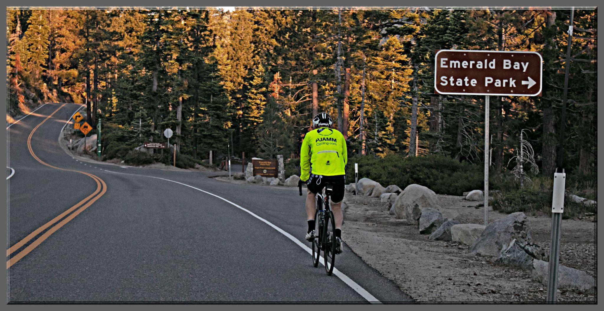 PJAMM Cyclist rides on two-lane mountain roadway surrounded by alpine forest; sign for Emerald Bay State Park