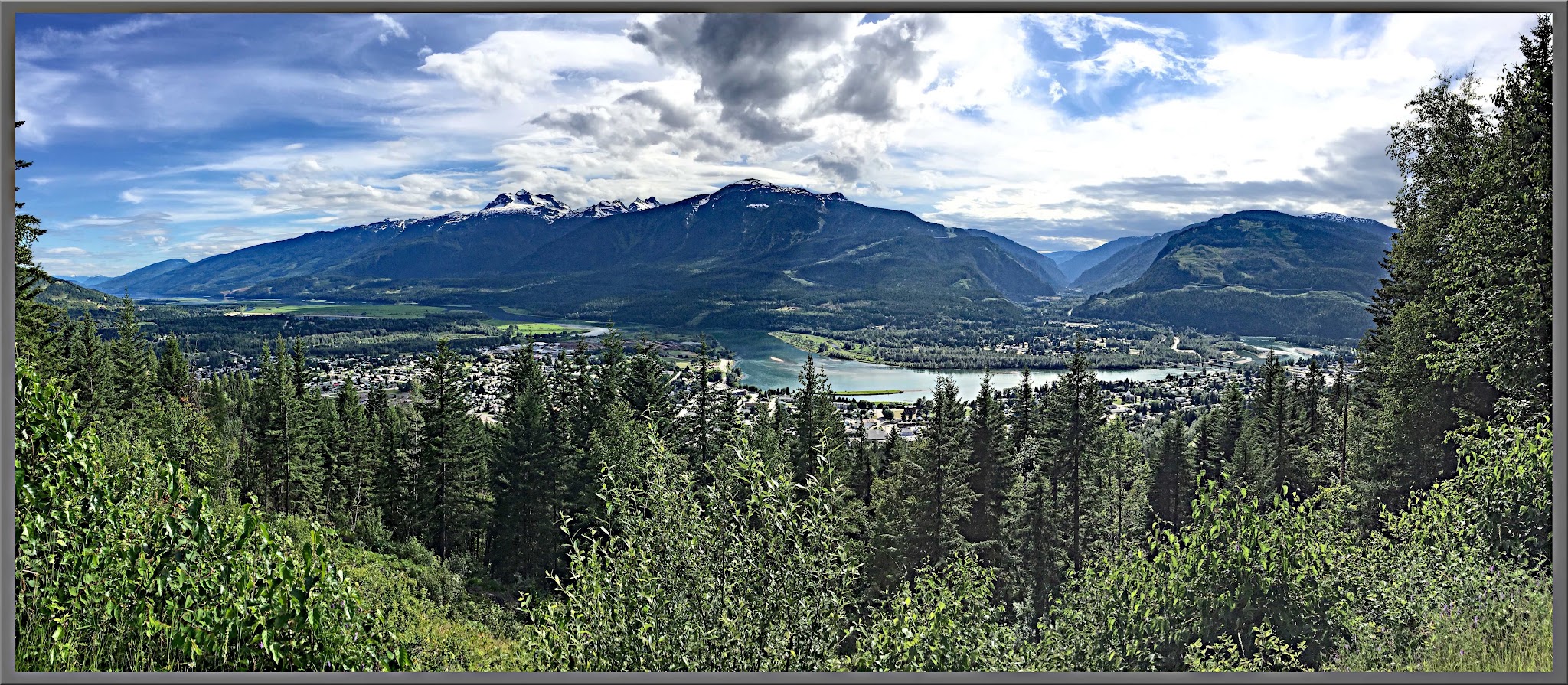 Cycling Mt. Revelstoke, BC, Canada - panoramic view of evergreen trees, a town in the distance, a large lake, and mountain range topped  in white clouds