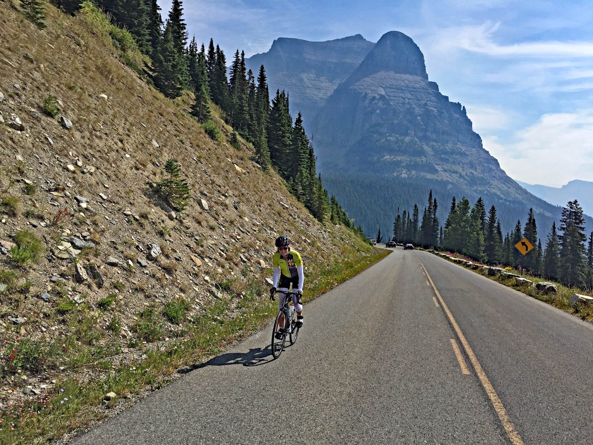 Cycling Logan Pass East, Going-to-the-Sun Road, Glacier National Park  - cyclist riding on straight portion of two-lane road with pointy mountain peaks behind