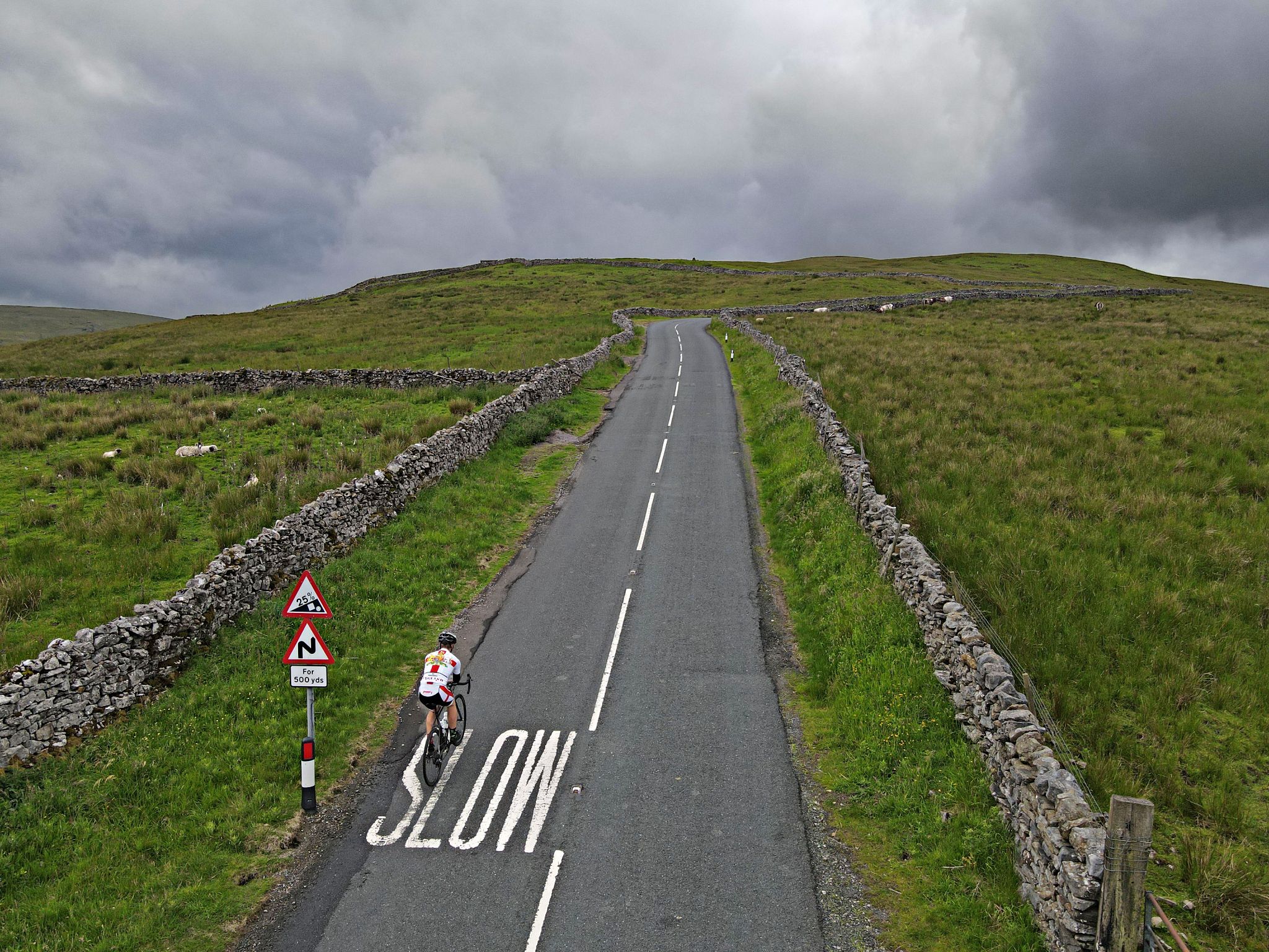 aerial view of PJAMM Cyclist climbing on two-lane roadway lined in stone walls surrounded by green pastureland