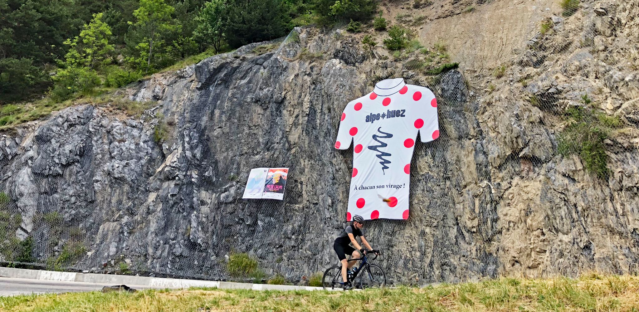 Tour de France 2023 cyclist rides by large polka dot jersey sign on rock wall, Alpe d'Huez