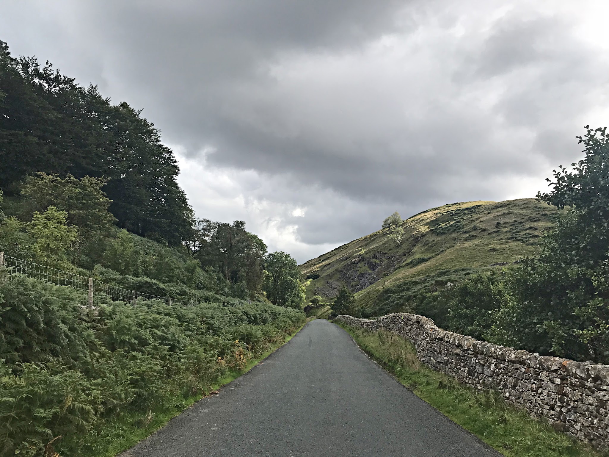 Cycling Trough of Bowland, Britain  - straight portion of road leading uphill, lush greenery on one side and stone fence along other, cloudy grey skies