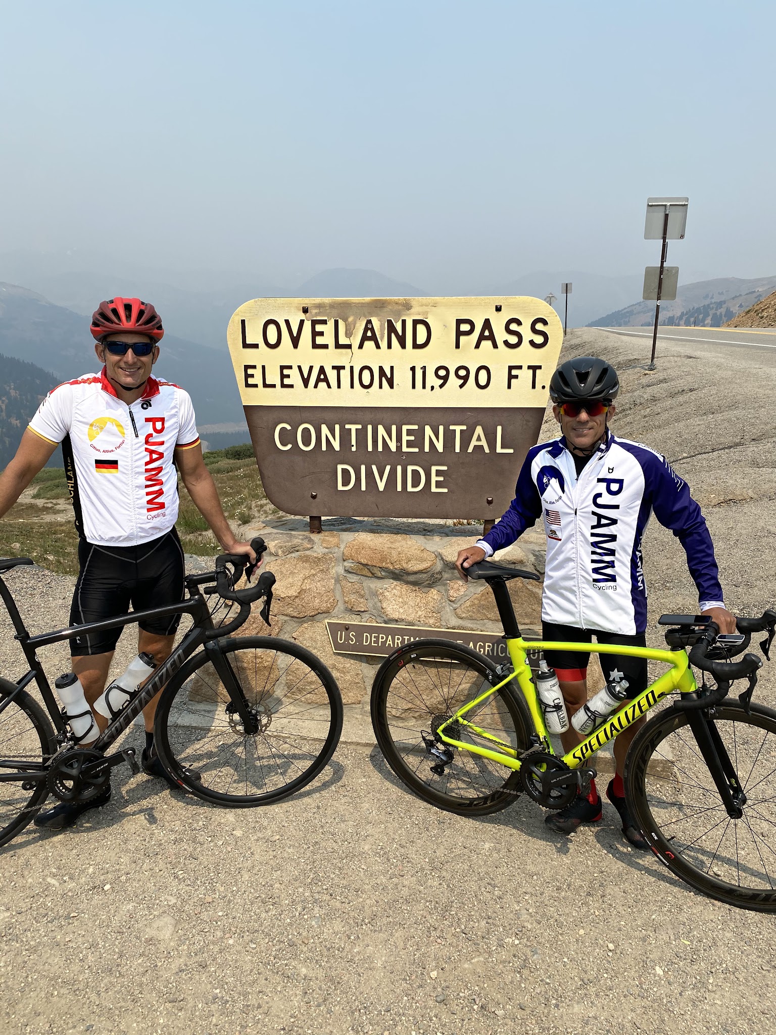 Cycling Loveland Pass from Georgetown - cyclists Eric Kuehl and Todd Short standing in front of Loveland Pass Continental Divide sign, wearing PJAMM Cycling jerseys