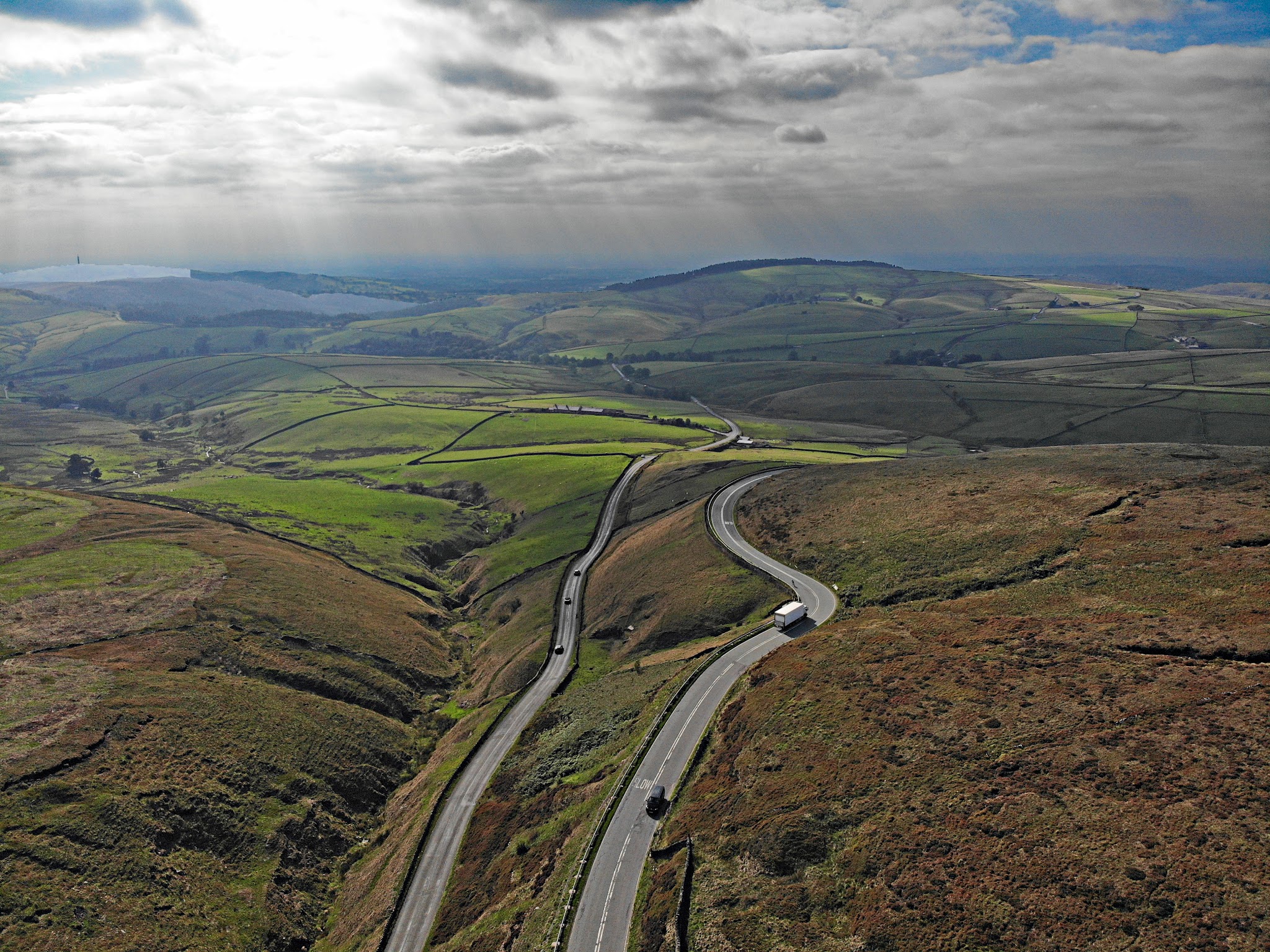 Cycling Cat & Fiddle, England - aerial view of long stretch of road stretching through pastureland, sunrays shining down through clouds