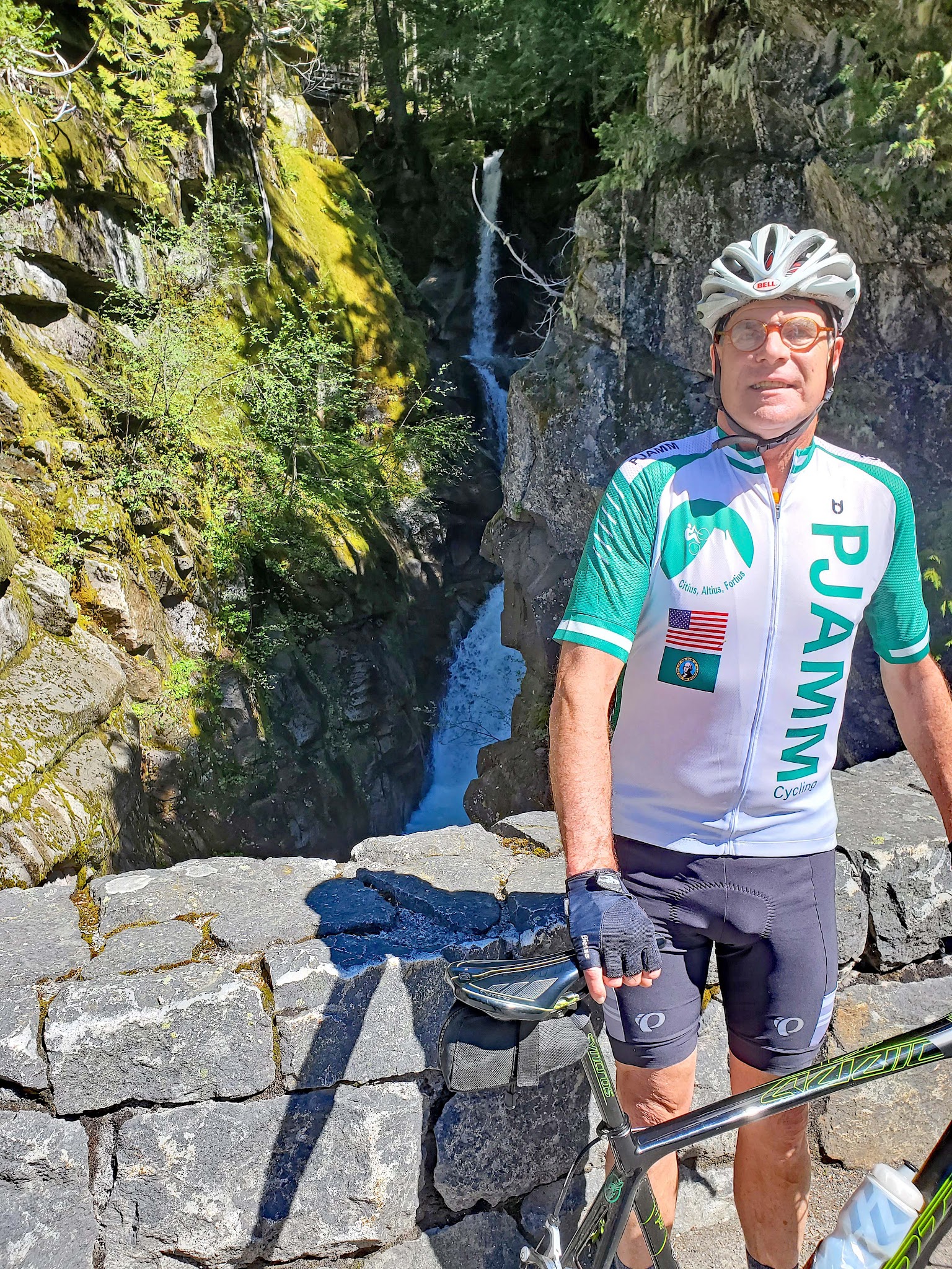 Cycling Mt. Rainier from Nisqually River Entrance - cyclist wearing white and green PJAMM Cycling jersey leans against low stone retaining wall in front of Christine Falls