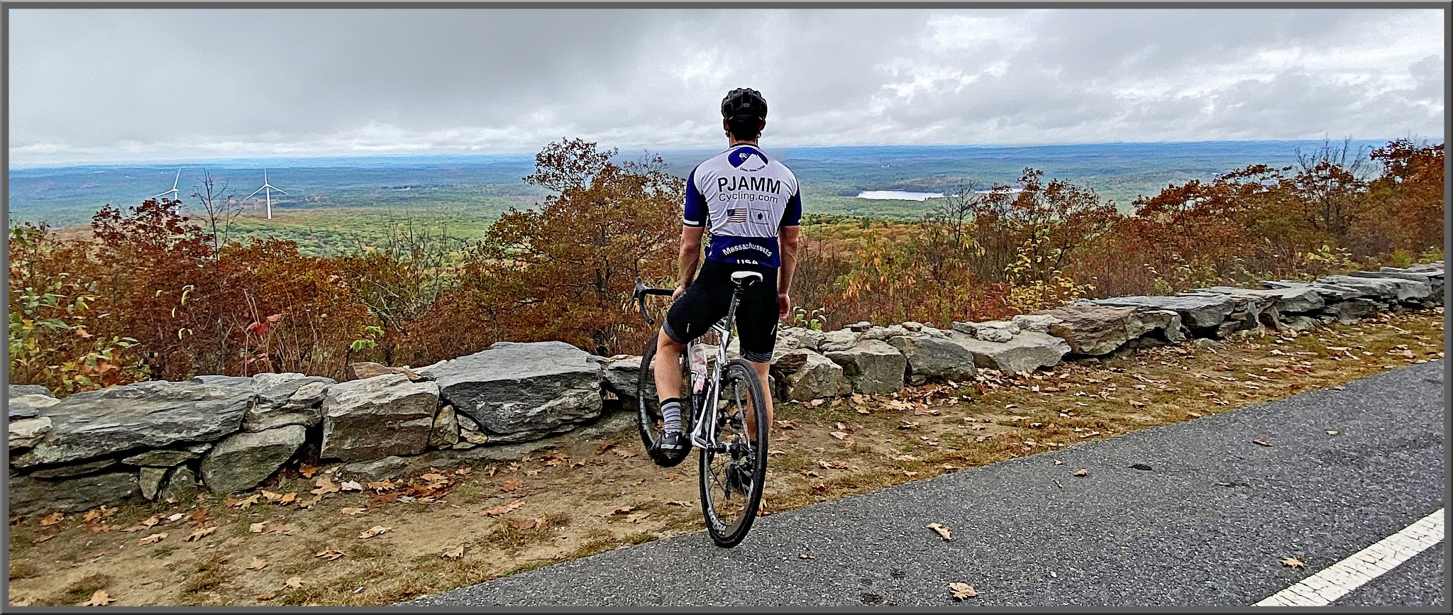 Cycling Wachusett Mountain North, Massachusetts  - PJAMM Cyclist stands on bike looking out over low retaining wall over fall foliage to greenery below