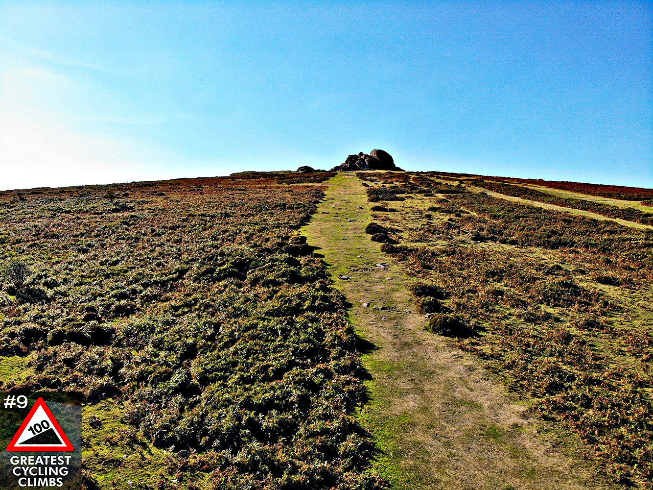 Cycling Haytor Vale - view from base of hillside looking directly up to summit, blue sky, rock formation at top, grass and shrubbery with a trail down center, Simon Warren's 100 Greatest Cycling Climbs #9 logo in corner