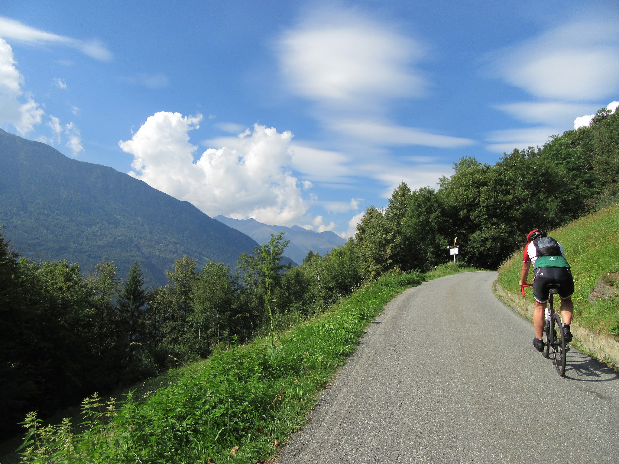Climbing Passo del Mortirolo from Mazzo di Valtellina by bike - cyclists riding on roadway with mountains and clouds in background