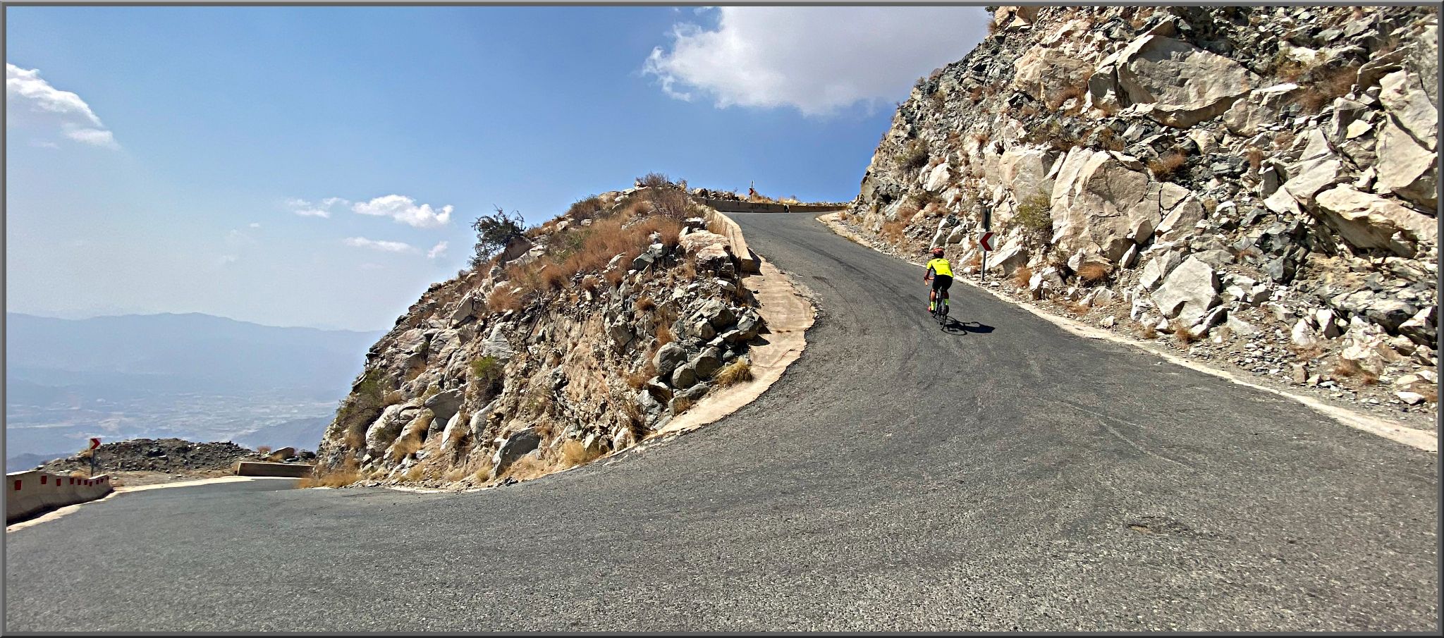 Cycling Baniamr Aquabah Pass, Saudi Arabia - PJAMM Cyclist rides up hillside beyond large hairpin turn, large rocks in landscape