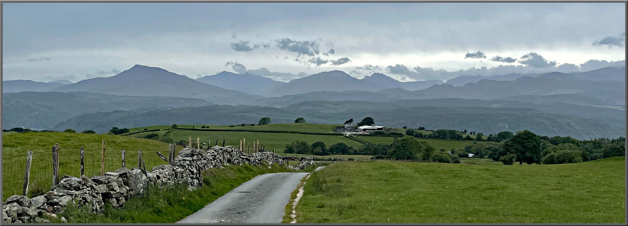 one-lane country road surrounded by green pastures; mountains in distance 