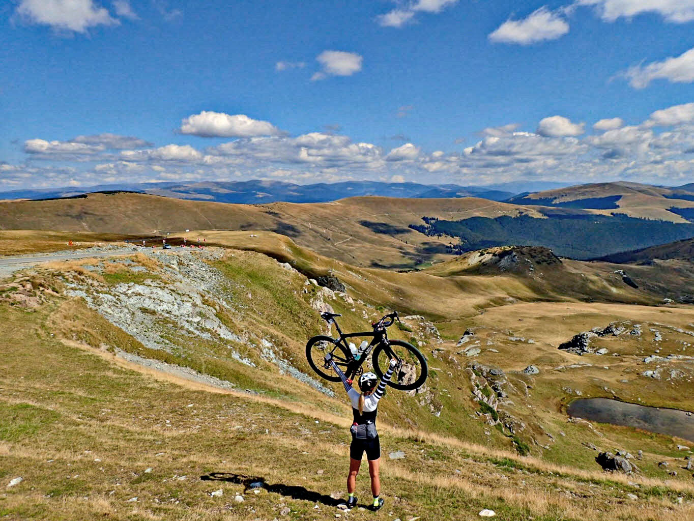 Cycling Transalpina North, Romania - cyclist stands on mountaintop overlooking hills below, holding her bike above her head