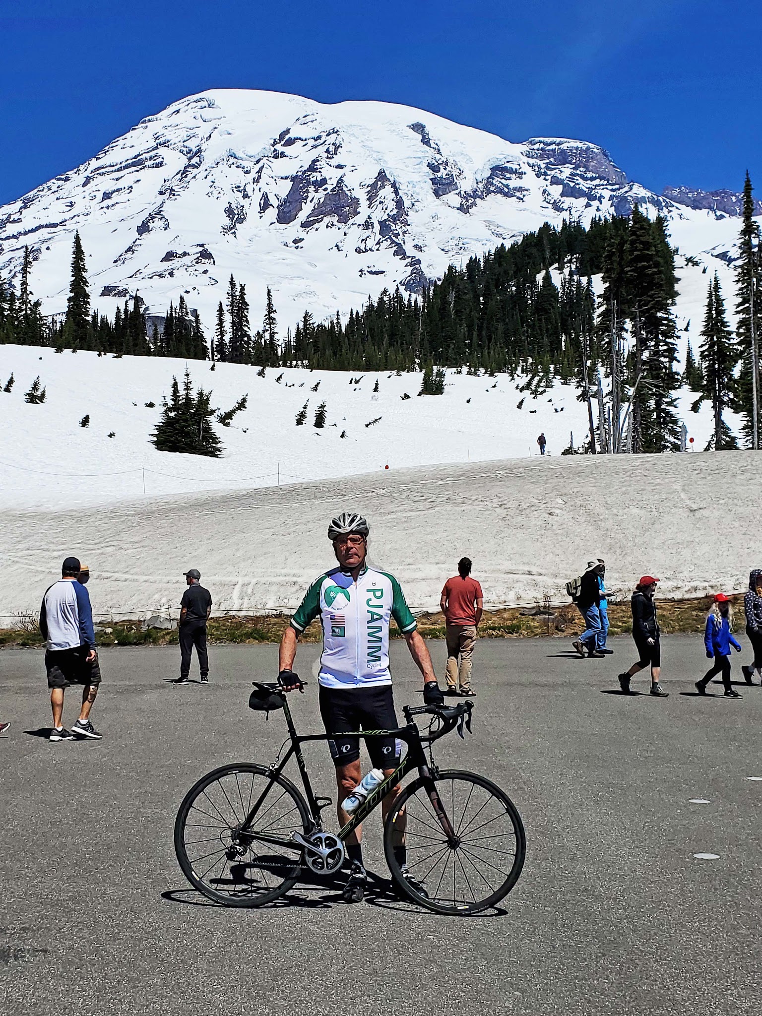 Cycling Mt. Rainier from Nisqually River Entrance - cyclist stands with bike at Paradise, snow capped mountain behind him