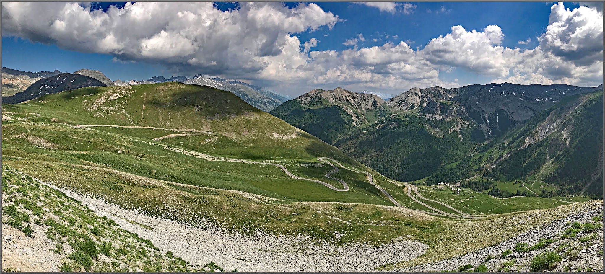 panoramic view shows roadside snaking and curving up mountain, green hillsides, blue sky with white clouds