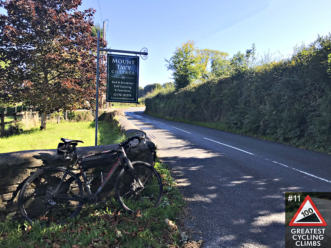 Cycling Rundlestone, England - road bike leaned against low rock wall next to hanging sign for Mount Tagvy Cottage along narrow two-lane road bordered by tall green hedges, Simon Warren's Greatest Cycling Climbs #11 logo in corner