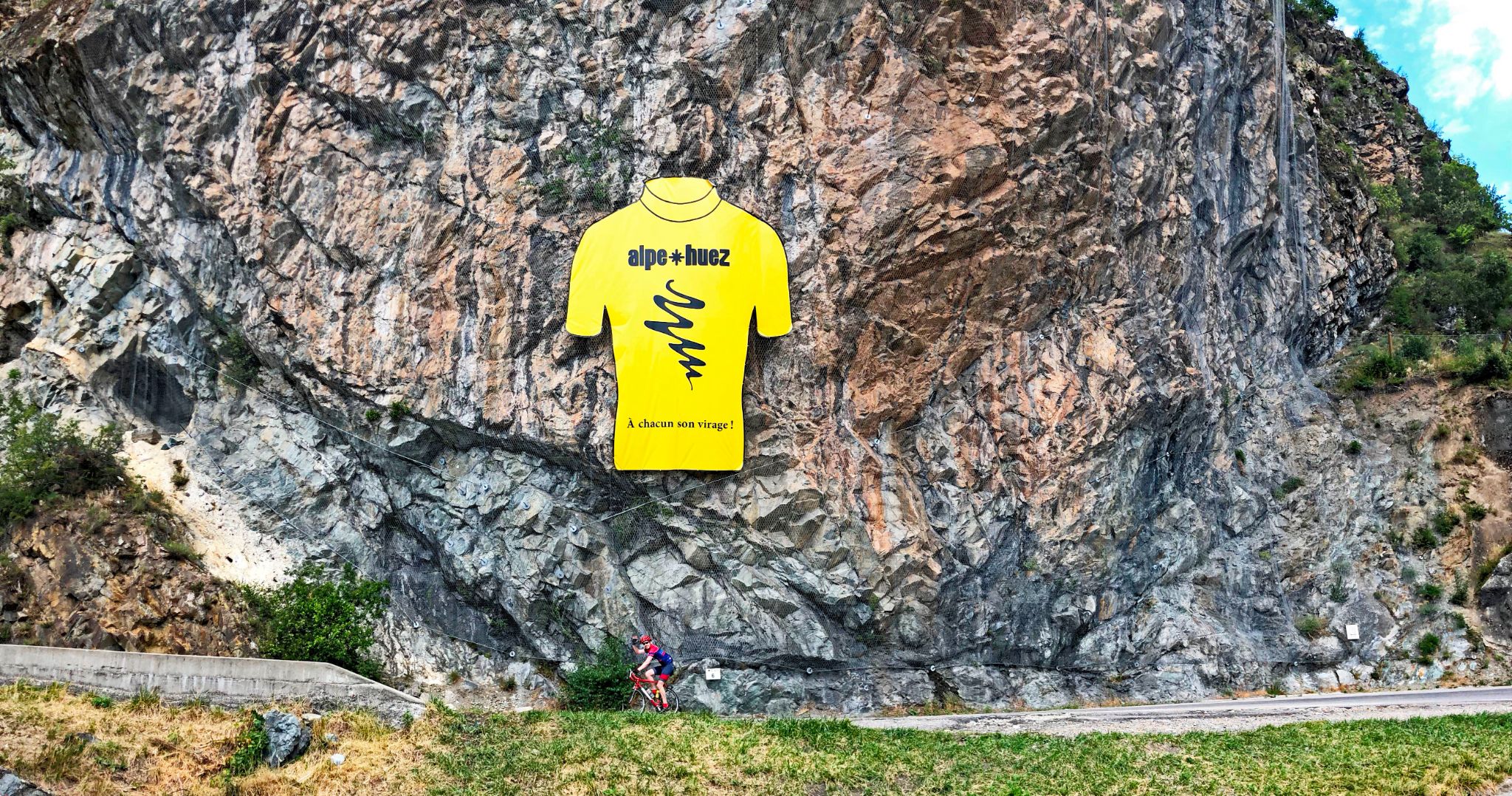 Alpe Huez, France - cyclist rides on roadway alongside huge rock wall with giant yellow jersey sign reading "Alpe Huez"