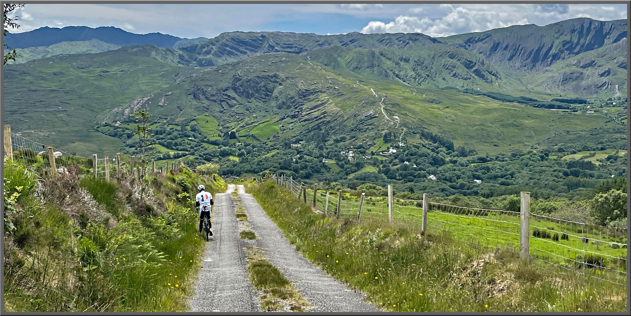 PJAMM Cyclist rides bike on one-lane narrow gravel country road, surrounded by green pastures and hills