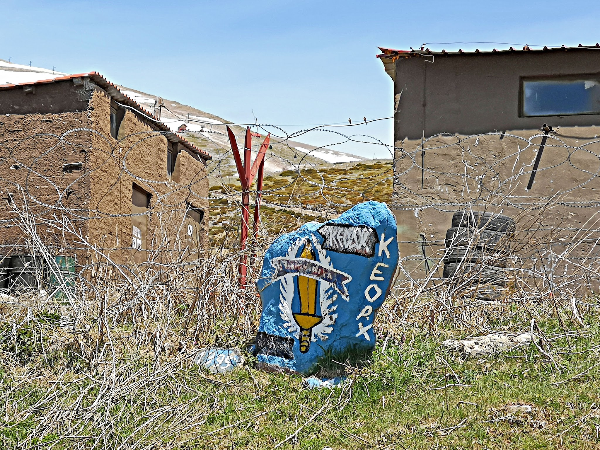 Cycling Mount Olympus, Greece - stone painted blue reading KEOAX with a yellow sword painted on it, in front of barbed wire with brown brick barracks-like buildings behind