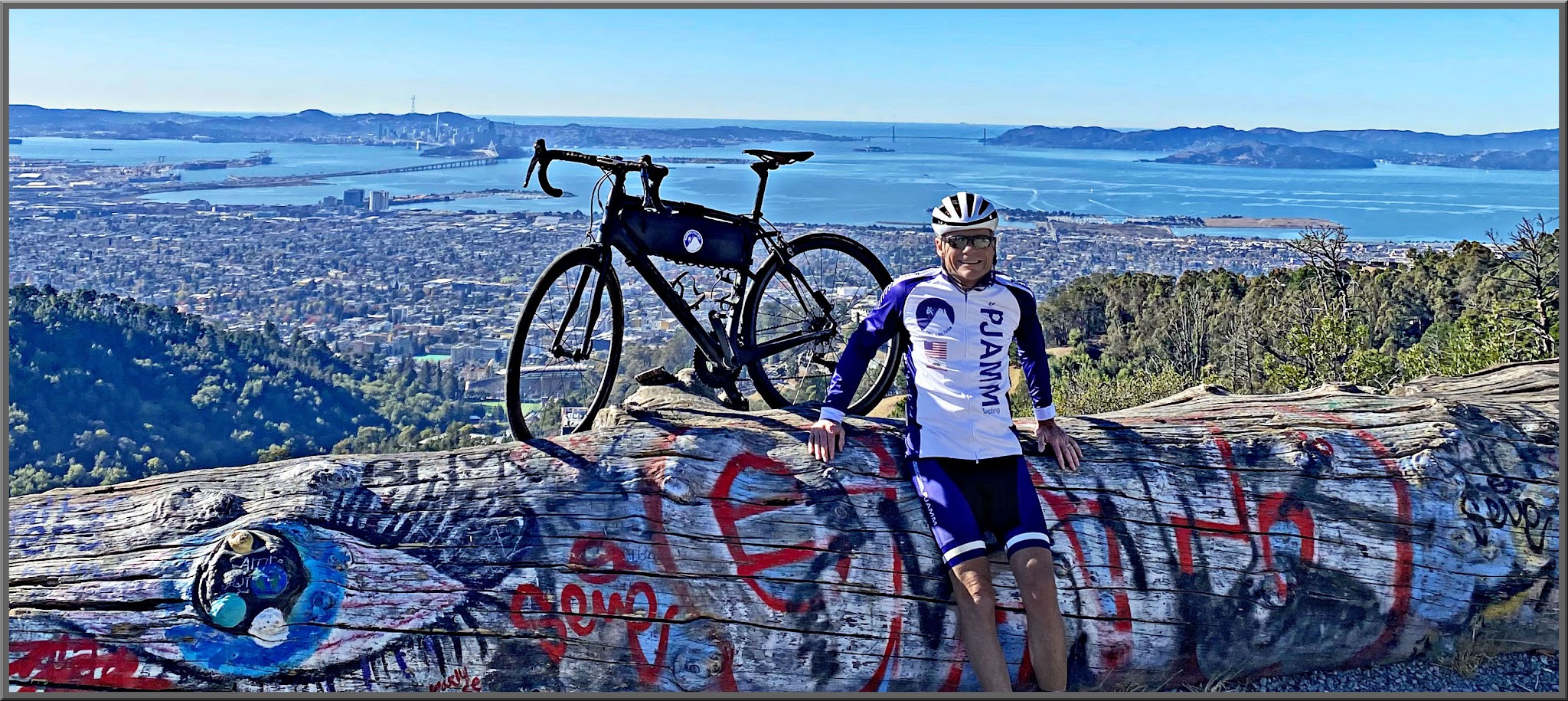 Cycling Tunnel to Grizzly Peak, California - PJAMM Cyclist John Johnson leads against large graffiti covered log with bike behind him, overlooking bay area and the bay beyond